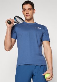 Blå bomull sport t-skjorte med korte ermer, rund hals og logo på brystet. Modellen holder en tennisrackett og en gul tennisball.