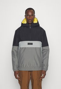 Chaqueta ligera con bloques de color que presenta una sección superior negra, un medio gris y un dobladillo elástico. Incluye una capucha forrada de amarillo y un parche con el logotipo.