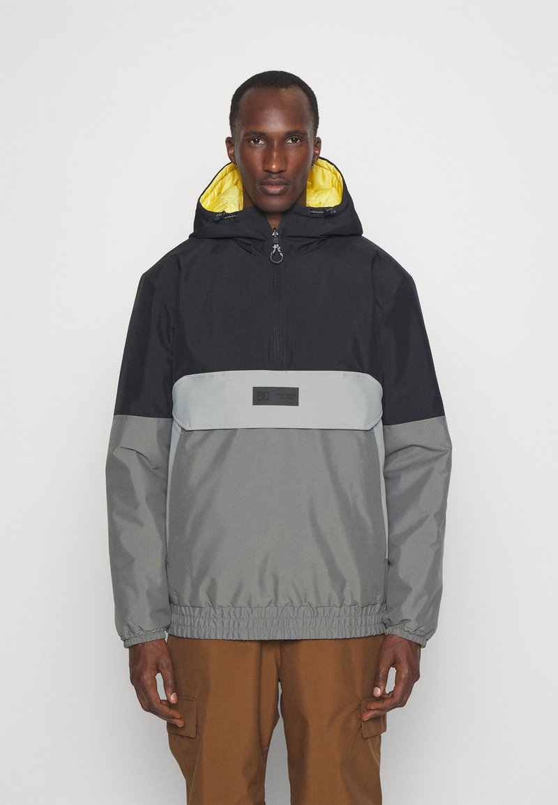 Chaqueta ligera con bloques de color que presenta una sección superior negra, un medio gris y un dobladillo elástico. Incluye una capucha forrada de amarillo y un parche con el logotipo.