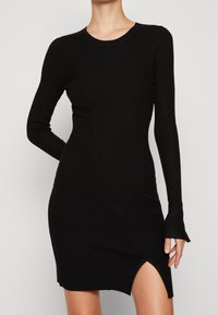 Robe noire côtelée à manches longues, col rond, fente latérale et coupe cintrée pour un look élégant. Texture douce avec tissu extensible.