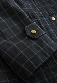 Jūrų laivyno plaid švarko detalė su auksiniu snap mygtuku, kontrastingomis siūlėmis, tekstūruota medžiaga ir kampuota kišenės uždanga.