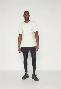 Muž stojící na jednobarevném pozadí, oblečený v bílém tričku Nike, černých šortkách přes černé legíny a černých sportovních botách.