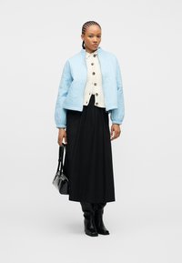 Γυναίκα με πλεγμένα μαλλιά που φοράει ανοιχτό μπλε καπιτονέ μπουφάν, κρεμ πουλόβερ με κουμπιά, μακριά μαύρη φούστα, μαύρες μπότες, κρατώντας μαύρη τσάντα.