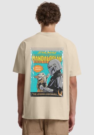 Personne portant un t-shirt beige avec une impression colorée de la couverture du comic Mandalorien montrant le Mandalorien tenant Baby Yoda en train de tendre la main.