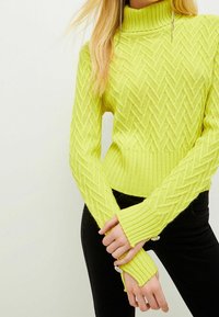 Yellow knitted sweater with a high collar, featuring a textured zigzag pattern and ribbed cuffs. Paired with black pants.