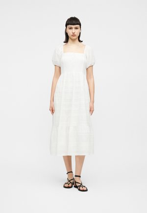 Młoda kobieta stoi, ubrana w białą midi sukienkę z bufkami na rękawach oraz czarne, płaskie sandały na paski, na jednolitym białym tle.