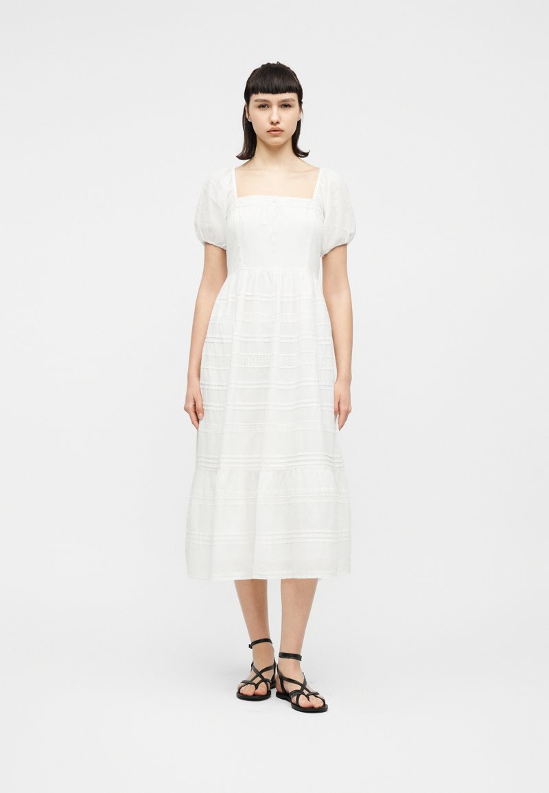 Νεαρή γυναίκα όρθια, φοράει λευκό μεσομπούστι φόρεμα με φουσκωτά μανίκια και μαύρα σανδάλια με λουράκια και επίπεδη σόλα, μπροστά σε απλό λευκό φόντο.