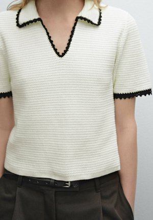 White short-sleeve knit top with black scalloped trim on collar and sleeves, worn with black pants and a black belt.