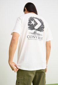 Camiseta de algodón blanca con un diseño gráfico negro de un árbol con una estrella y el texto "CONVERSE", cuello redondo, mangas cortas y corte relajado.