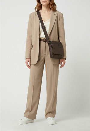 Una mujer luce un traje beige con zapatillas deportivas blancas y un bolso bandolera de color marrón oscuro, sobre un fondo de estudio liso.