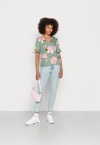 Top floral verde com mangas curtas e em babados; combinado com jeans azul claro e tênis brancos; a carregar uma pequena bolsa rosa.