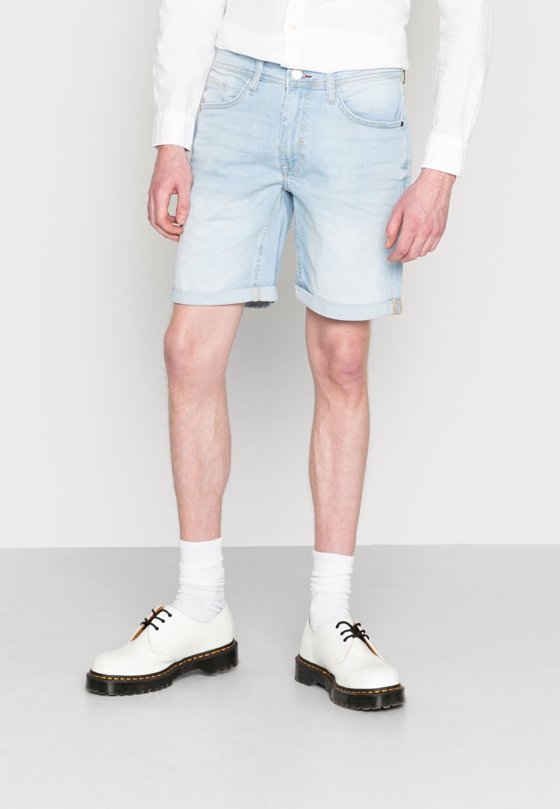 Shorts en denim bleu clair avec ourlets retroussés, associés à des chaussures blanches et des chaussettes. Présente un design classique à cinq poches et une légère décoloration.