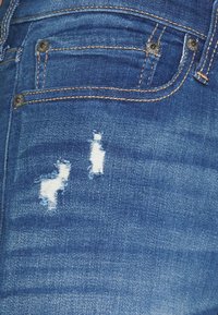 Dunkelblaue Denim-Jeans mit einer verblassten Textur, ausgestattet mit zwei abgenutzten Stellen und metallischem silbernen Zubehör in der Nähe der Taschen.