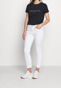 Žena oblečená v černém tričku s logem Tommy Hilfiger, bílých slim-fit džínách a bílých teniskách, stojící na neutrálním světlem pozadí.