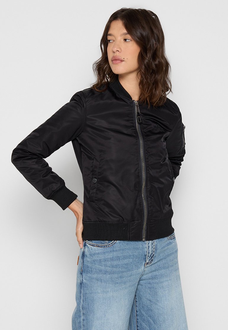 Czarna kurtka bomberka z zamkiem błyskawicznym z przodu, ściągaczami na mankietach i u dołu, gładka w dotyku, z bocznymi kieszeniami, zestawiona z jasnoniebieskimi dżinsami.