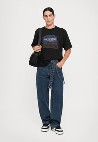 Camiseta negra con texto gráfico en azul y marrón, combinada con jeans azules holgados y un bolso cruzado negro. Zapatillas en blanco y negro.