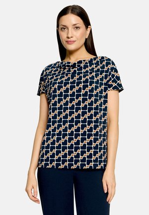 Femme aux cheveux bruns raides, portant un haut bleu marine à manches courtes avec un motif géométrique et un pantalon foncé sur un fond uni.
