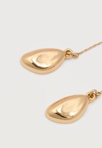 Δύο γυαλιστερά χρυσά μενταγιόν σε σχήμα δακρύου κρέμονται από λεπτές χρυσές αλυσίδες σε λευκό φόντο.