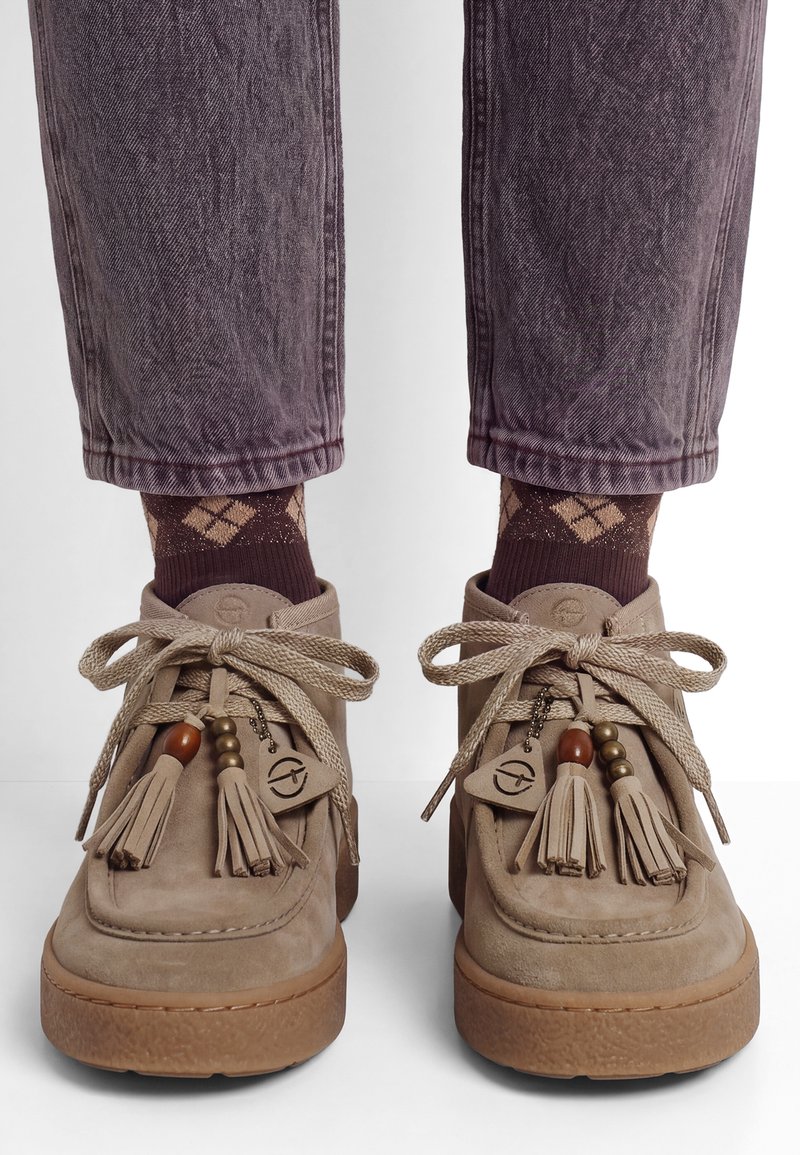Chaussures en daim beige avec décorations de glands et perles, lacets noués, portées avec des chaussettes marron à motif losanges et un pantalon violet foncé retroussé.
