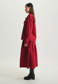 Chemise en coton rouge avec un col pointu, associée à une jupe rouge à volants. La robe présente des manches longues et des accents de boutons. Bottines noires en cuir. 