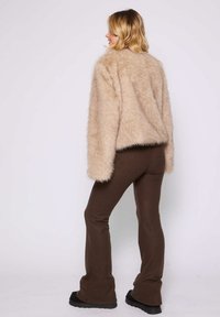 Beige Kunstfelljacke mit lockerer Passform, langen Ärmeln und cropped Saum. Kombiniert mit braunen Bootcut-Hosen und schwarzen flachen Schuhen.