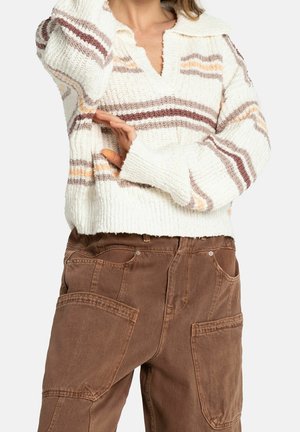 Donna che indossa un maglione bianco a maglia con strisce marroni e pesca e pantaloni marroni a vita alta con grandi tasche frontali.