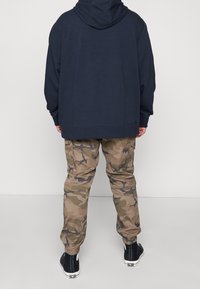 Henkilö pukeutuneena tummansiniseen huppariin, ruskean ja vihreän maastokuvioisiin cargohousuihin, joissa on resorit nilkoissa, ja mustiin korkeavartisiin lenkkareihin, seisomassa valkoista taustaa vasten.