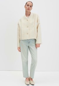 Vrouw in een crèmekleurige textuur-gebuttonte jack, lichtblauwe jeans en beige instapschoenen, staand tegen een witte achtergrond.