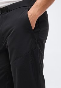 Άνδρας με μαύρο παντελόνι, με το χέρι στην πλαϊνή τσέπη, με γωνιακό ραφές και διακριτικό λογότυπο της μάρκας στον μηρό.