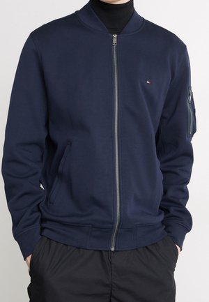 Mężczyzna ubrany w granatową bomberkę zapinaną na zamek, z małym logo na piersi i zapinanym na zamek kieszonką na lewym rękawie, ręce w kieszeniach.