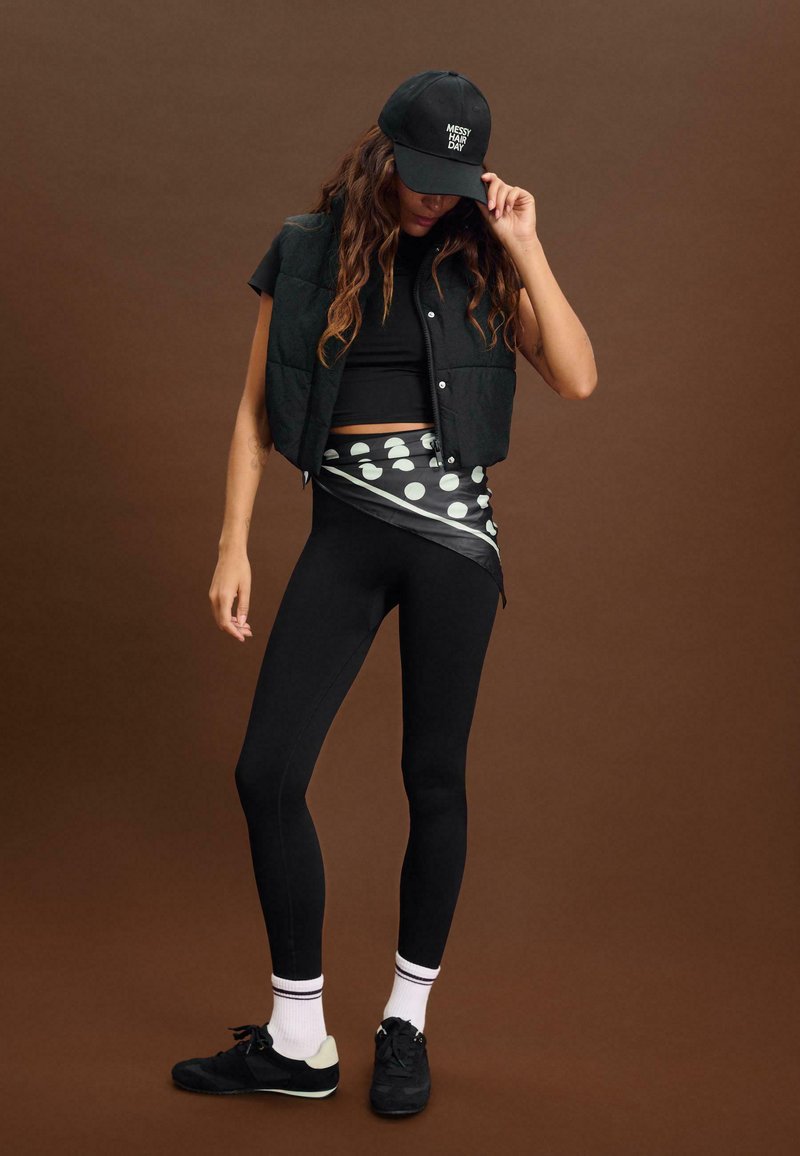 Schwarze, voluminöse Weste über einem schwarzen Crop-Top, schwarzen Leggings und schwarzen Turnschuhen. Akzentuiert mit einem grau-weißen gepunkteten Tuch und einer schwarzen Mütze.