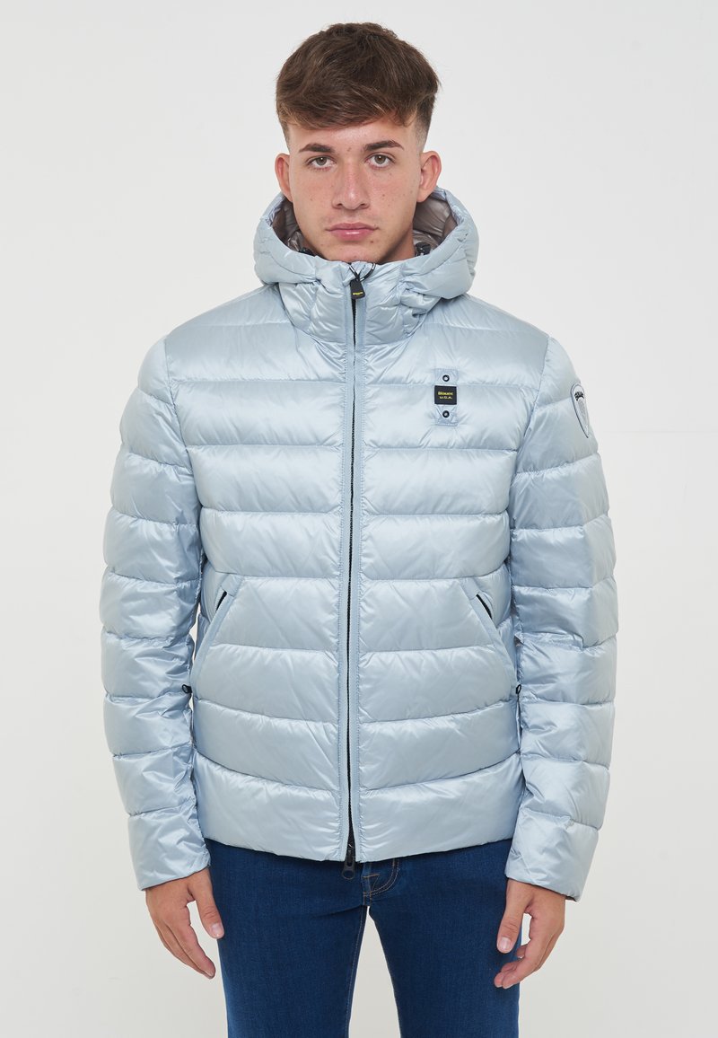 Giacca isolante blu chiaro con finitura lucida, cappuccio, zip frontale e due tasche laterali. Presenta pannelli trapuntati orizzontali.