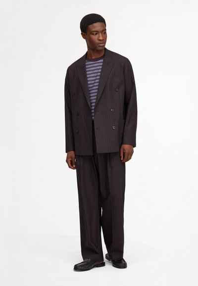 Man wearing loose dark pinstripe suit with double-breasted jacket over a striped shirt and black leather shoes standing against white background.