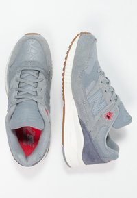 Grijze sportieve schoenen met een mix van suede en mesh materialen. Kenmerken zijn een rubberen zool, een gevoerde tong en rode accenten op de binnenkant en het logo.