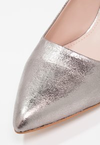 Pompe argentée métallisée à bout pointu avec une surface texturée, présentant un design à découpe basse et un intérieur beige. Semelle plate et finition lisse.