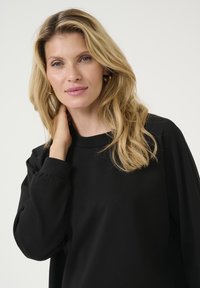 Crna majica dugih rukava s okruglim izrezom i napuhanim rukavima, izrađena od mekog materijala. Glatka tekstura i opušteni kroj s detaljima na manžetama.