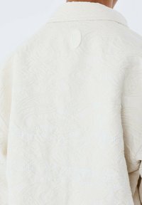 Krämfärgad jacka med en präglad blommönster, vit sömnad och en rund logotagg vid kragen. Texturerad tygdetalj synlig.