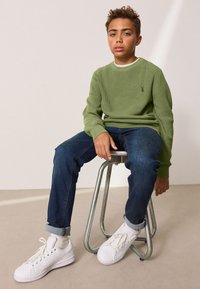 Vihreä ribbineulepaita, siniset farkut ja valkoiset tennarit. Istuu metallijakkaralla vaaleaa taustaa vasten. Rentoon tyyliin, kerroksittain pukeutunut.