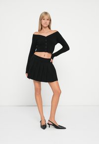 Crna pletena kratka jakna s gumbima, uparena s crnom suknjom s nabranim detaljima. Model nosi crne slingback potpetice, stoji nasuprot bijele pozadine.