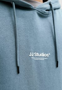 Felpa con cappuccio azzurro chiaro con coulisse e logo bianco "JJ Studios". Tessuto morbido, design casual, con dettagli neri sulle coulisse.