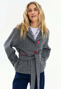 Žena s dlhými blond vlasmi nosí čierno-biely pásikavý kabát s ružovými gombíkmi a tmavomodré nohavice na jednoliatom pozadí.