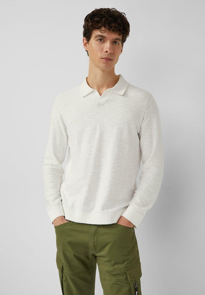 Camisa polo blanca de manga larga con cuello, tejido de punto texturizado y puños ajustados, combinada con pantalones cargo verdes con bolsillos laterales.