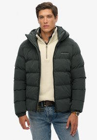 Chaqueta de plumas verde oscuro con capucha, cuenta con paneles cosidos y cierre de cremallera. Llevada sobre un suéter beige claro, combinada con jeans azules.
