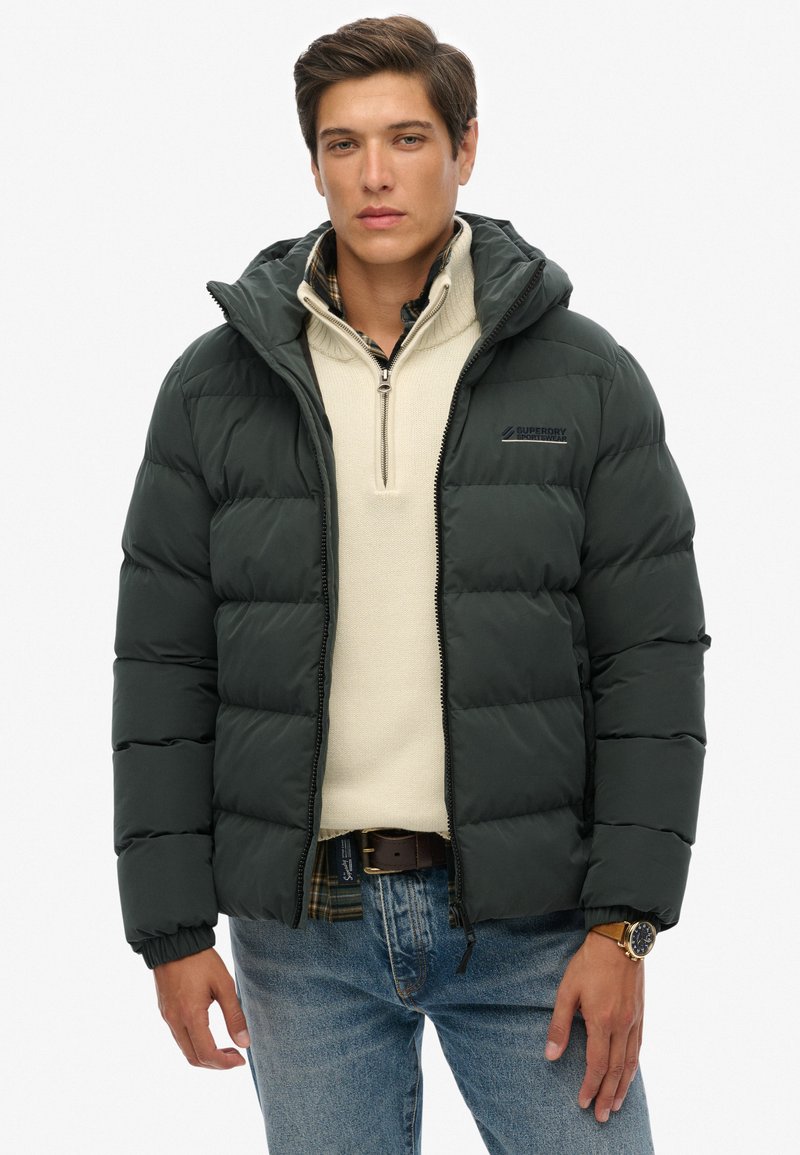 Chaqueta de plumas verde oscuro con capucha, cuenta con paneles cosidos y cierre de cremallera. Llevada sobre un suéter beige claro, combinada con jeans azules.