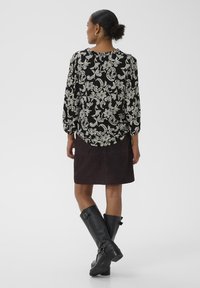 Frau steht mit dem Rücken zur Kamera, trägt eine schwarze Bluse mit floralem Muster, einen dunklen Rock und knielange schwarze Lederstiefel vor einem weißen Hintergrund.