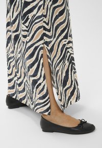 Ben der bærer sorte ballerina-flade sko og en zebra-stribet nederdel med slids forneden, der viser underben og ankel, på hvid baggrund.