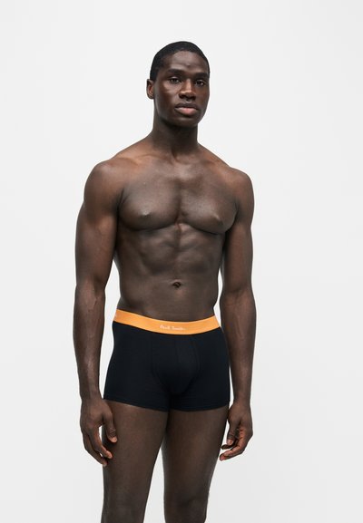 Homme musclé debout devant un fond uni, portant un boxer noir avec une ceinture orange et le logo Paul Smith.