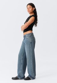 Woman with long curly hair wearing a black sleeveless top, wide-leg blue jeans, black shoes, standing with arms crossed against white background.