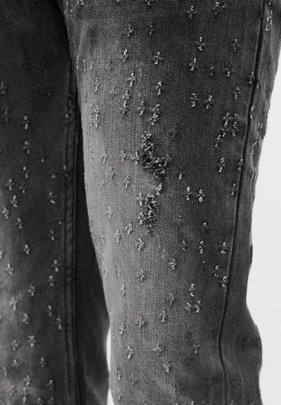 Κοντινό πλάνο μαύρου τζιν με ξεφτισμένες, ανάγλυφες κουκκίδες διασκορπισμένες πάνω στο ύφασμα, που αναδεικνύουν τις λεπτομέρειες του φθαρμένου σχεδίου.