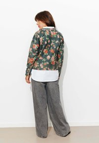 Cardigan vert à motif floral avec manches longues, poignets côtelés, et une chemise blanche à col en dessous, assortis à un pantalon gris à jambes larges.